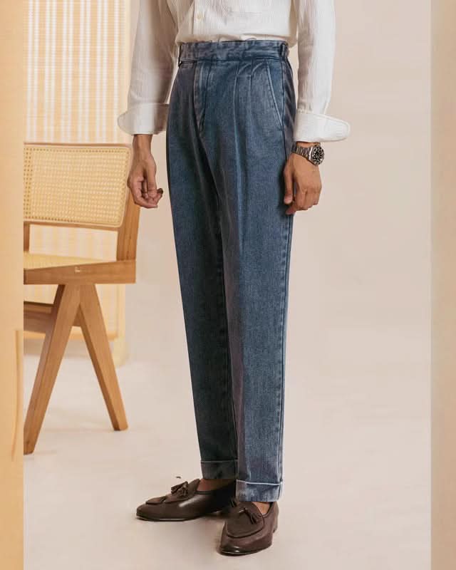 A person stands indoors wearing Balfour Ateliers Pisa Pleated Denim Trousers, styled with a white long-sleeve shirt and brown tassel loafers. A woven-back wooden chair appears behind; the persons face remains unseen.