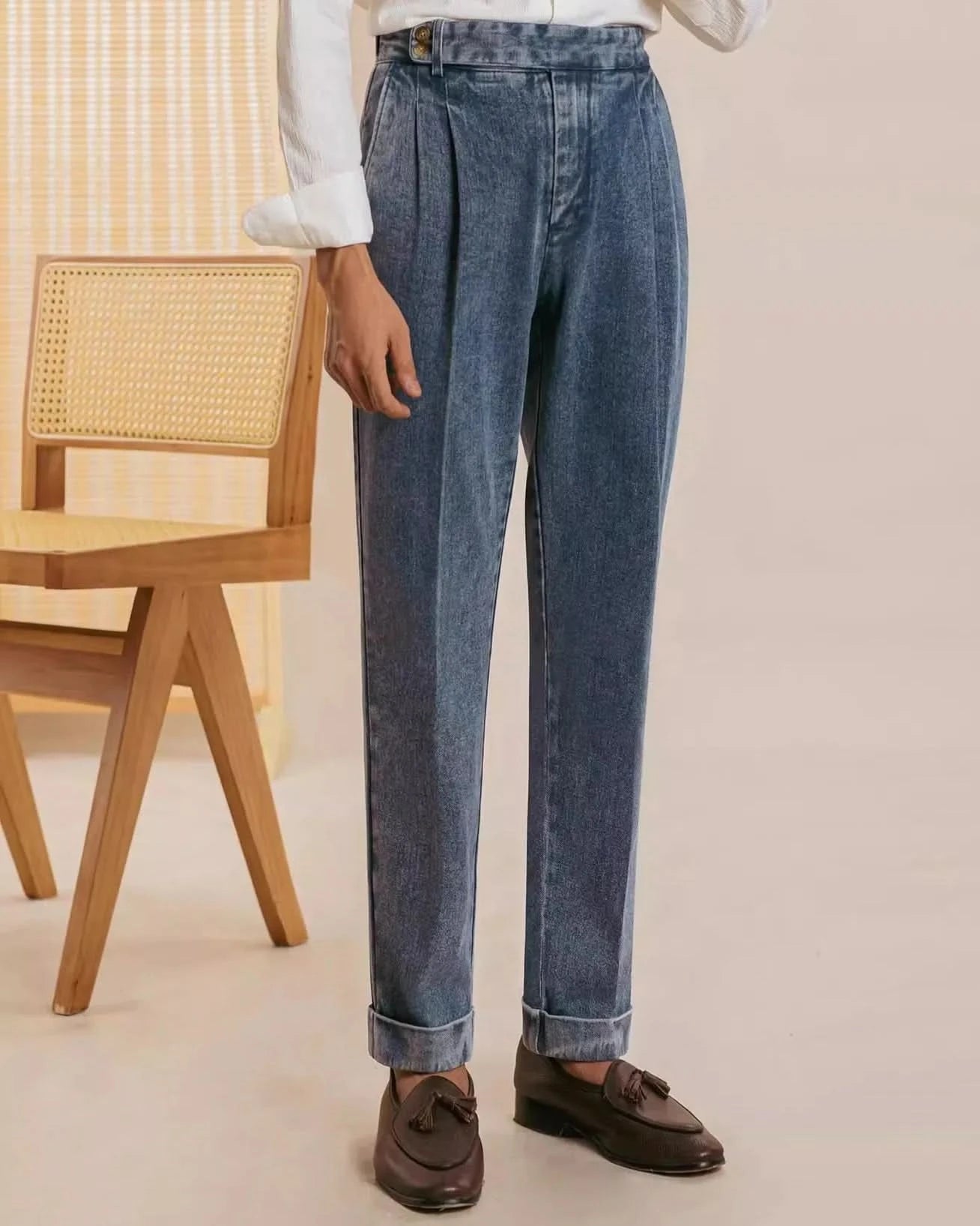 Indoors, a person wears Balfour Ateliers Pisa Pleated Denim Trousers—high-waisted with rolled cuffs—paired with a white long-sleeve shirt and dark brown tassel loafers. Italian-inspired tailoring shines. A cane-back wooden chair is visible behind them.