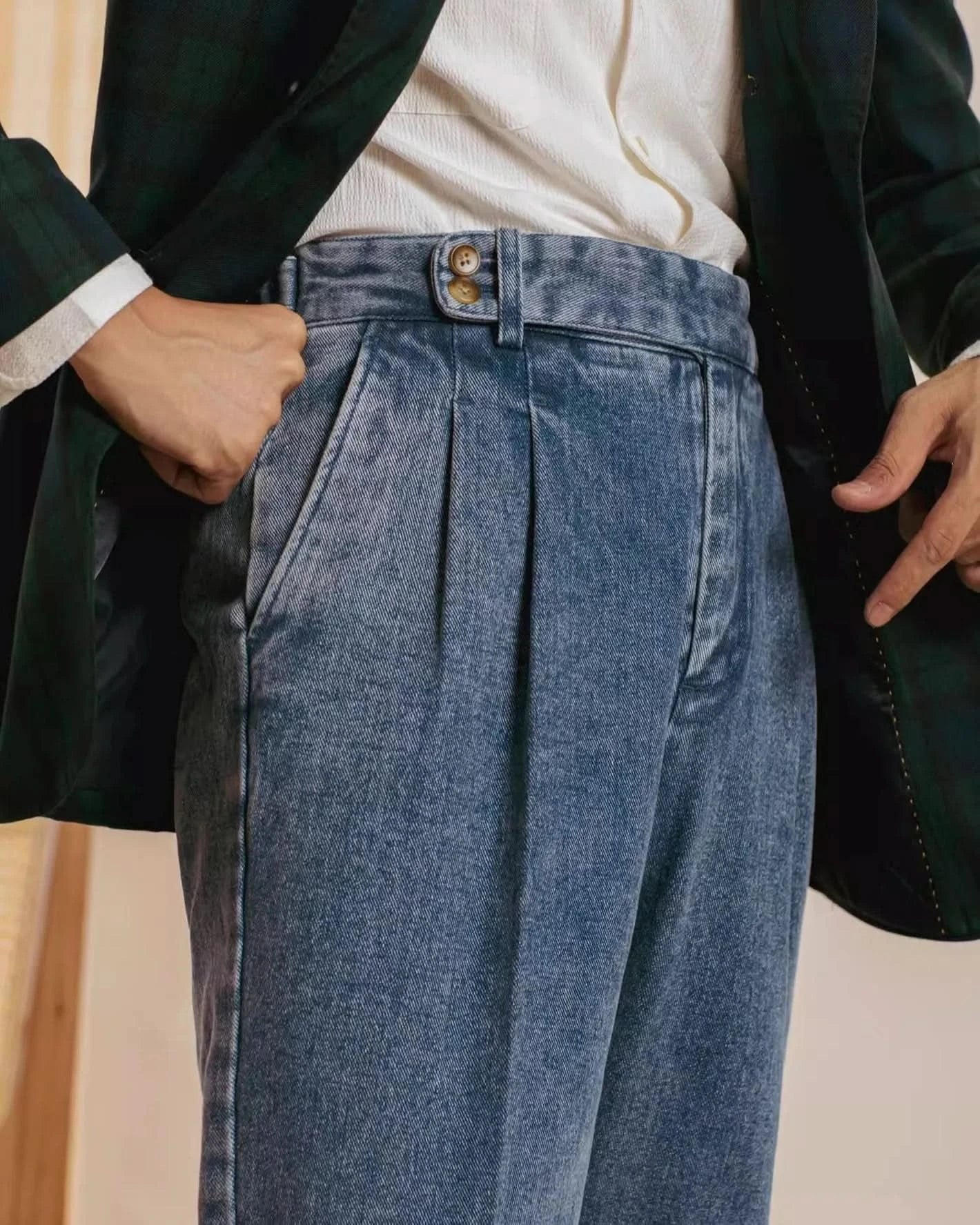 A person wears Balfour Atelier’s Pisa Pleated Denim Trousers with a white textured shirt tucked in and a dark green plaid blazer. One hand in the pocket, the other pointing to the trousers to highlight their Italian-inspired tailoring.