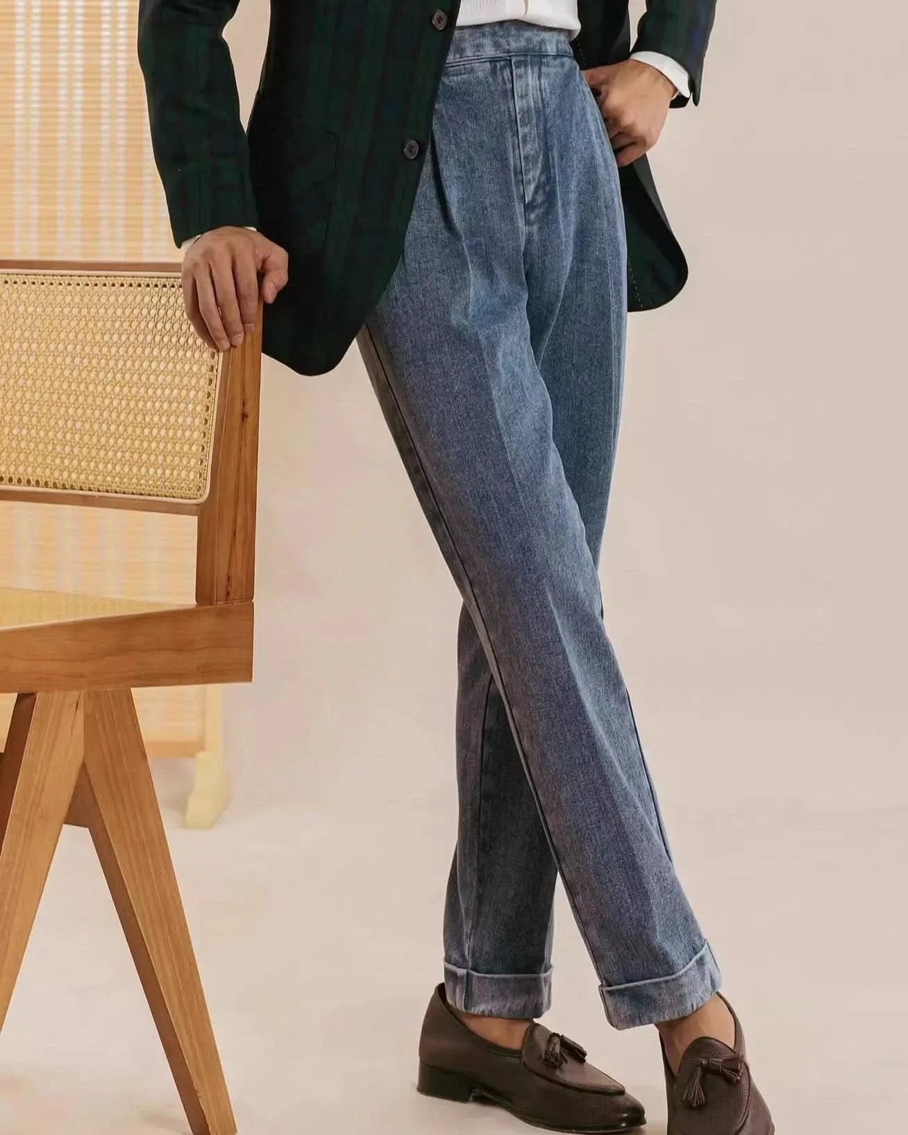 Indoors, a person leans on a wooden cane-back chair, wearing Balfour Atelier’s Pisa Pleated Denim Trousers styled as mens tailored pants, with a dark green plaid blazer, white shirt, and brown tassel loafers.