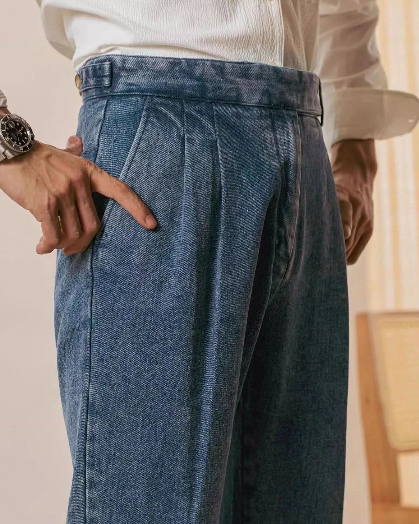 A person wears Balfour Atelier’s Pisa Pleated Denim Trousers with a white shirt, pointing to the front pocket. The Italian-inspired look is complemented by a wristwatch and a softly lit wooden chair in the background.