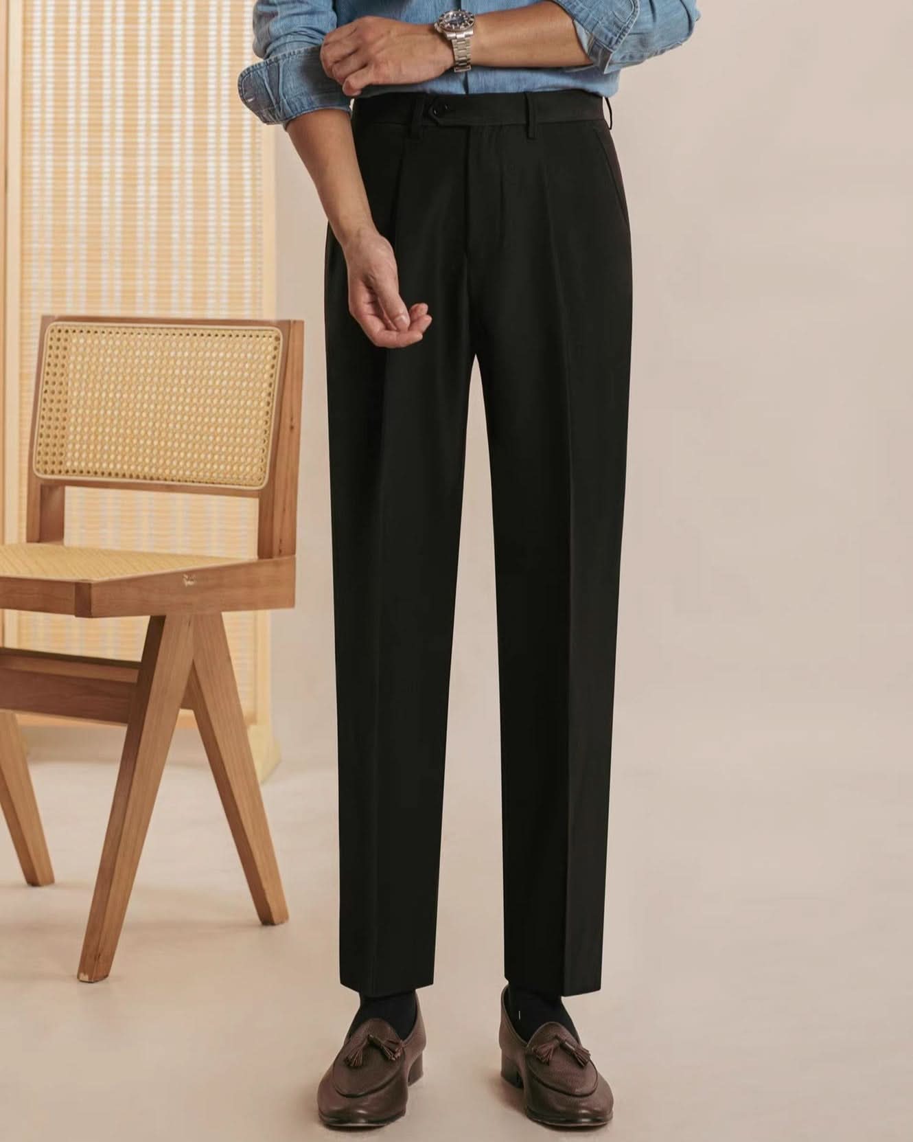 A person wears Orta Tailored Trousers – Four Seasons Elegance by CjDropshipping, styled with a light blue button-up shirt and brown loafers. Their face is unseen. A wooden chair with a cane back is positioned to the left against a light background.