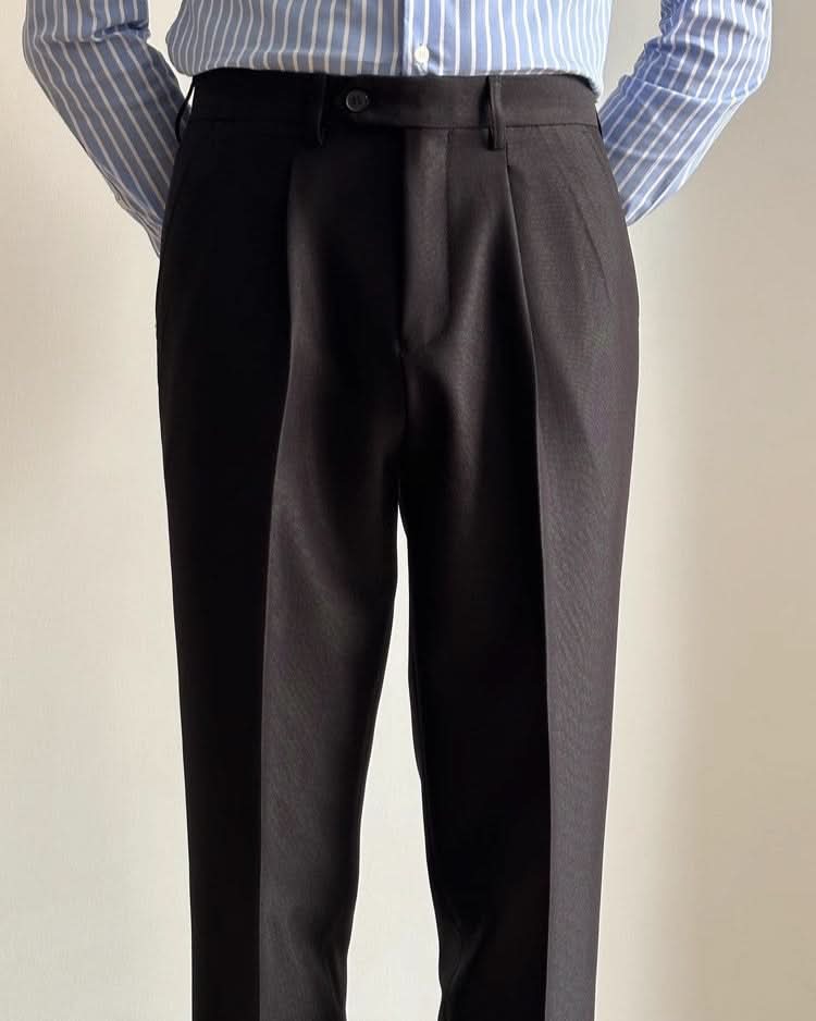 A person stands with hands behind their back, wearing CjDropshippings Orta Tailored Trousers – Four Seasons Elegance in black and a blue-white striped shirt tucked in, showcasing casual sophistication from waist to mid-calf against a plain light background.