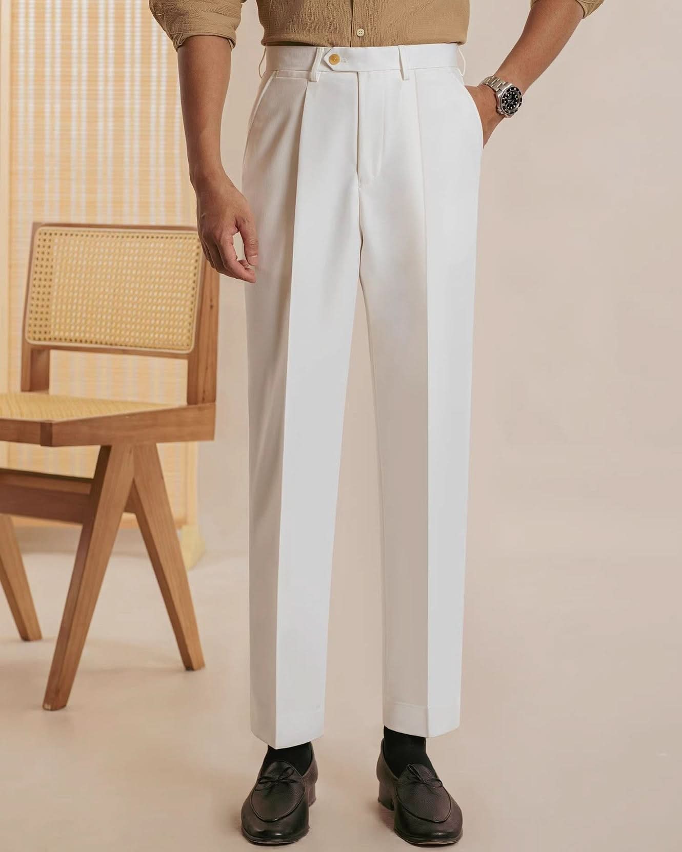 A person models the Orta Tailored Trousers – Four Seasons Elegance by CjDropshipping in white, styled with a tucked-in beige shirt, wristwatch, and dark loafers beside a wooden cane-back chair against a neutral indoor background. The head is not visible.