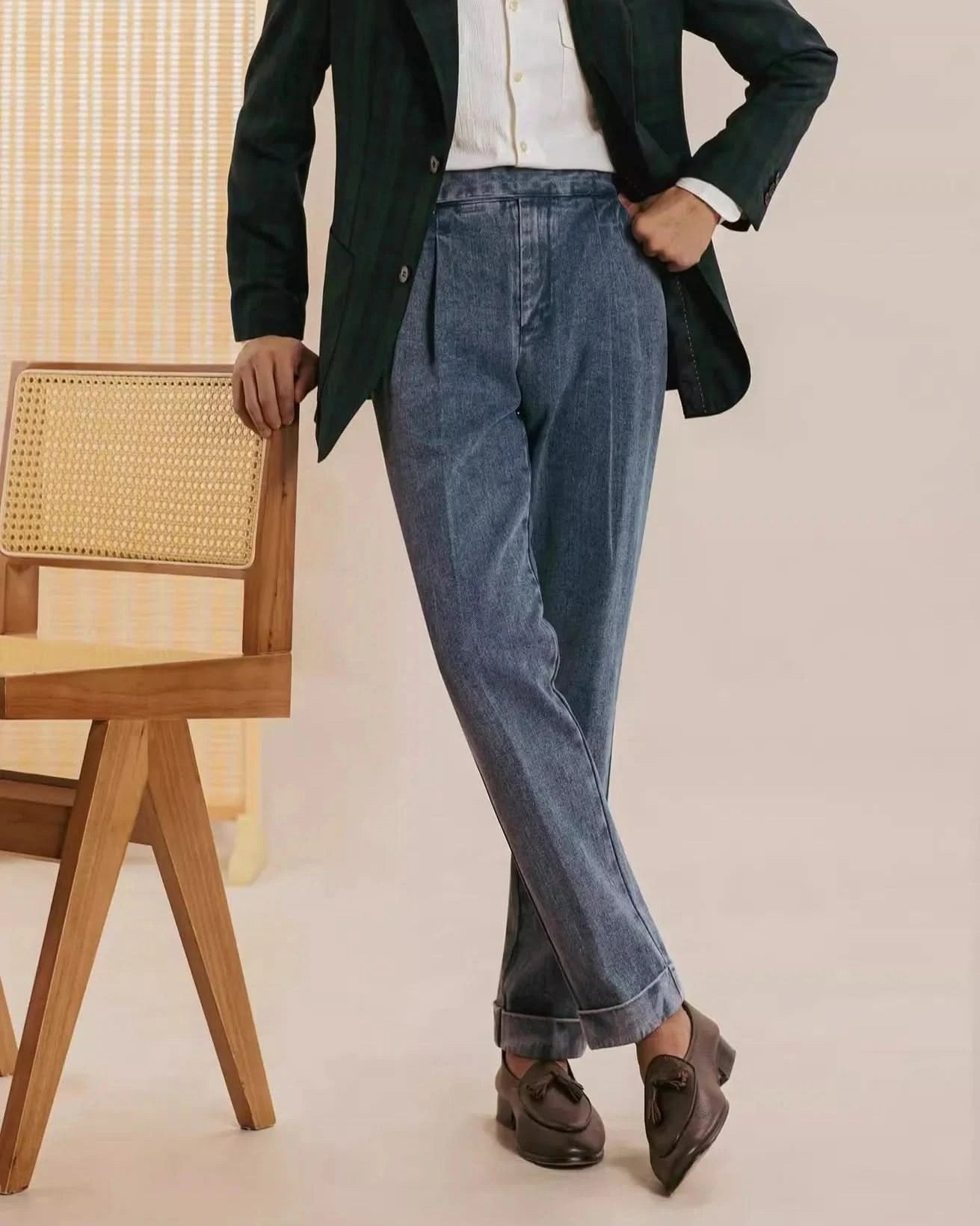 A person stands with legs crossed, leaning on a wooden chair, wearing Balfour Atelier’s Pisa Pleated Denim Trousers. Styled with a white shirt, green blazer, and brown loafers, they pose against a neutral backdrop; their head is not visible.